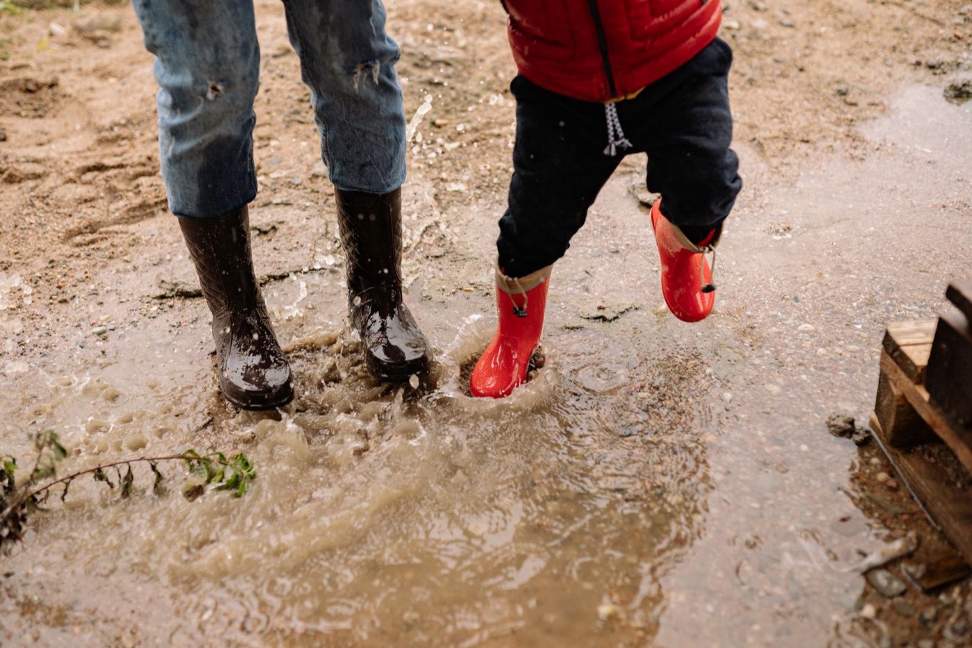 Two kids in rain boots splashing in a muddy puddle
